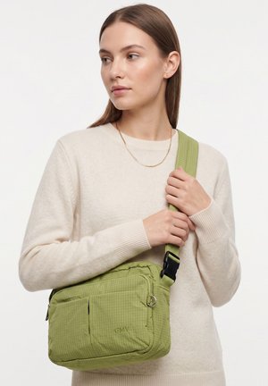 Giovane donna con maglione beige che tiene una borsa a tracolla verde con tracolla regolabile su sfondo bianco.