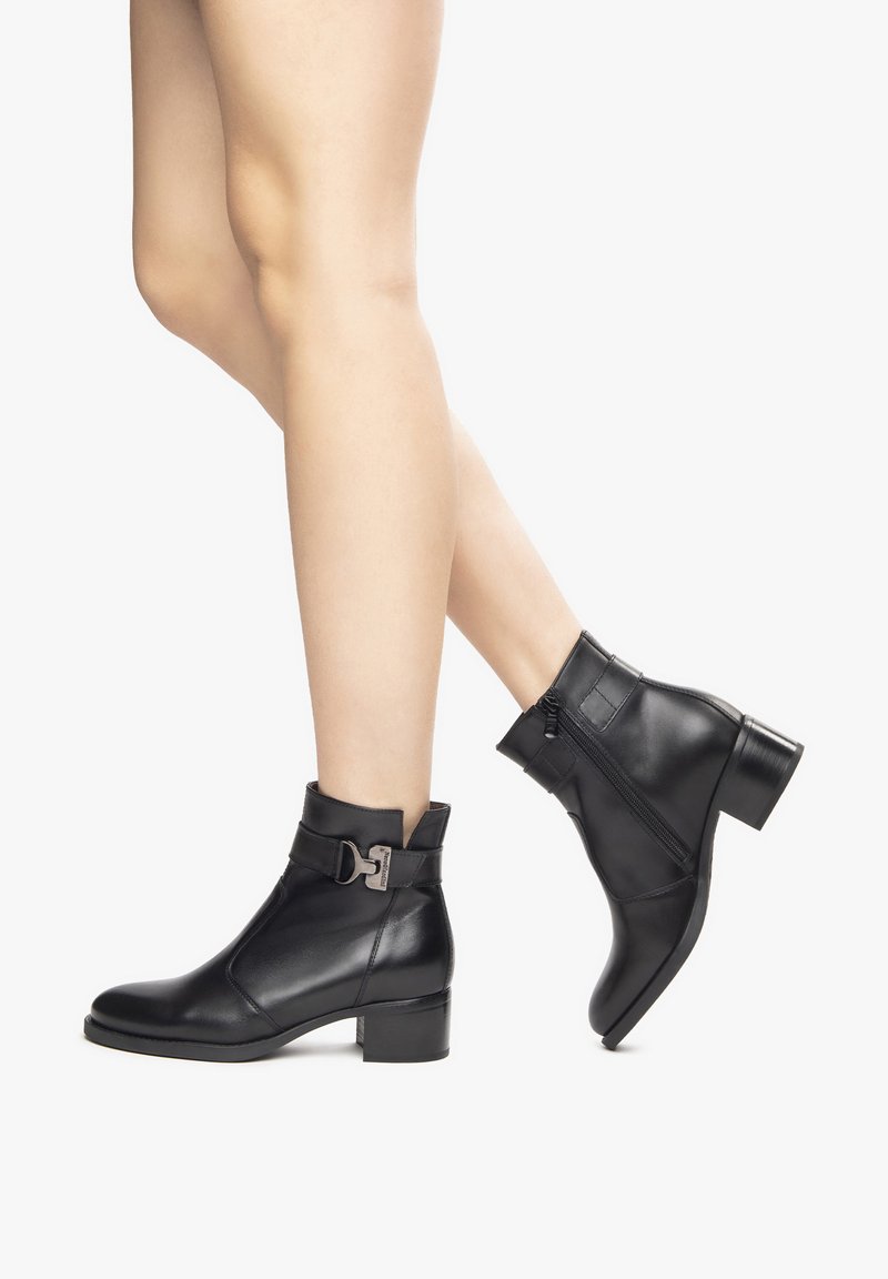 Bottines en cuir noir avec un petit talon, détails de sangle à boucle et fermeture éclair latérale pour un enfilage facile. Texture lisse, design épuré.