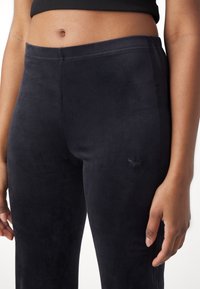 Black velour leggings with a smooth texture, featuring a subtle logo on the left side and side stripes in a slightly darker shade.