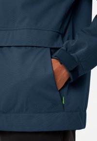 Veste imperméable marine avec une poche zippée, des détails cousus et un logo brodé. Texture lisse avec des poignets ajustables et un design peplum.