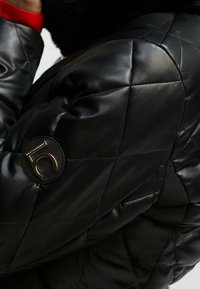 Quiltad svart skinnjacka med en cirkulär logotypmärke på ärmen. Har en slät textur och ett kontrasterande rött innerfoder.