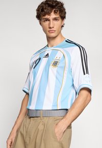 Φανέλα ποδοσφαίρου Αργεντινής σε ανοιχτό γαλάζιο και λευκές ρίγες, κοντά μανίκια, με μαύρα λογότυπα και λεπτομέρειες Adidas. Διαθέτει σήμα στο στήθος.