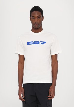 T-shirt en coton blanc avec col rond, manches courtes, arborant un logo EA7 bleu imprimé sur le devant. Design épuré, coupe classique.