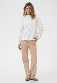 Blouse en coton blanc avec un col volanté et des détails en dentelle, associée à un pantalon en velours côtelé rose clair. Modèle portant des chaussures plates avec des clous.