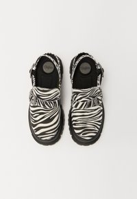 Zebra print klompe v črno-belem odtenku, s teksturiranim zgornjim delom, prilagodljivo naramnico in gumijastim podplatom. Okrogla konica in oblazinjen notranji obsod.
