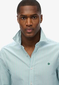 Camisa de botones a rayas azul claro y blanca con un cuello y un logo bordado verde en el pecho. Textura de tela suave.