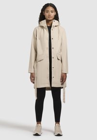 Trench coat impermeabile beige con cappuccio, bottoni a pressione, tasche laterali e vita regolabile con laccetto. Indossato sopra un top nero e leggings.