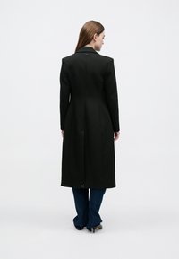 Hosszú fekete kabát testre szabott szabással, hajtónyakú gallérral, sima textúrával és finom hátsó részlettel. Sötét farmer fölött viselve.