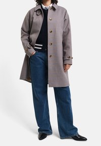 Brun og hvid ternet trench coat med store knapper, krave og sidelommer, parret med blå vidde jeans og sorte spidse sko.