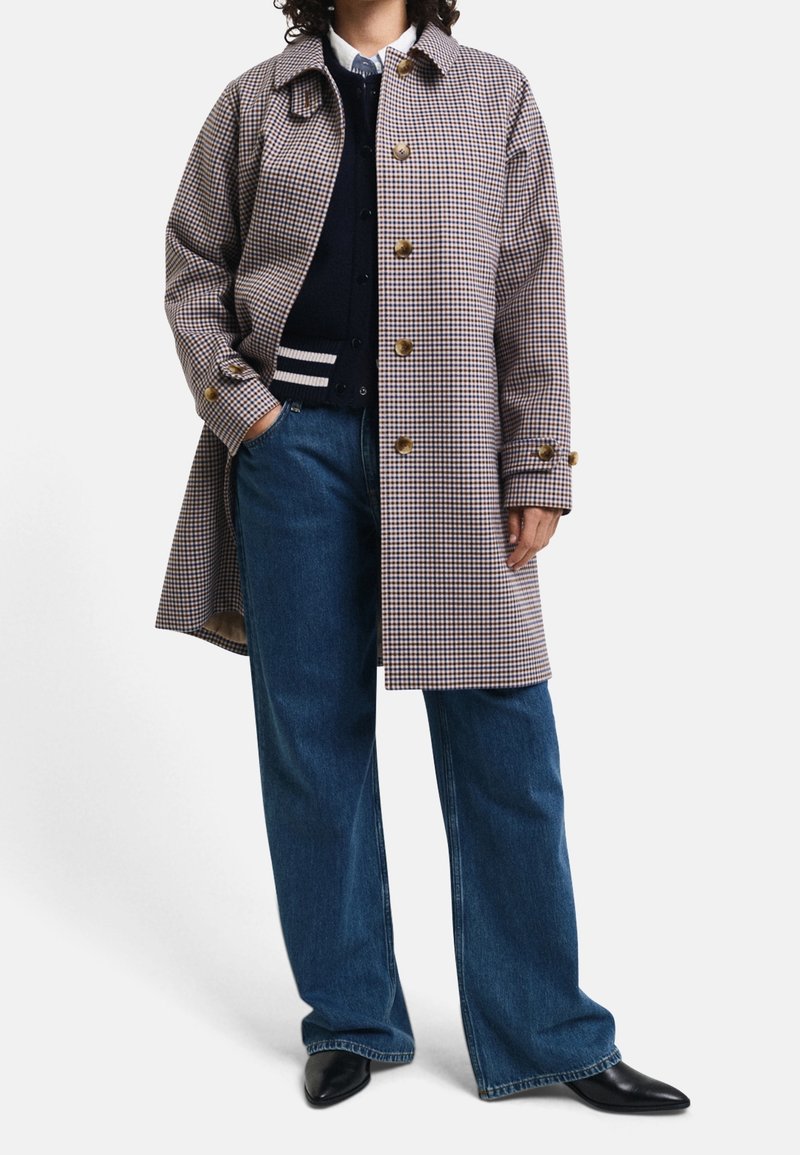 Brun og hvid ternet trench coat med store knapper, krave og sidelommer, parret med blå vidde jeans og sorte spidse sko.