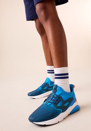Lábak fehér zokniban, kék csíkokkal és kék sportcipőben, fehér talppal világos háttér előtt.