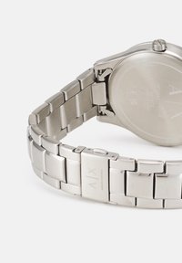 Srebrna ura iz nehrjavečega jekla s povezanim zapestjem, ki ima polirano in matirano površino. Na hrbtni strani je vgravirano logotip blagovne znamke in podrobnosti.