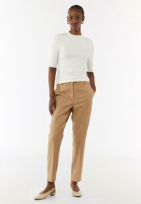 Biały top z krótkim rękawem w połączeniu z beżowymi spodniami o dopasowanym kroju i cielistymi sandałami na obcasie. Modelka stoi na tle jednolitej białej ściany.