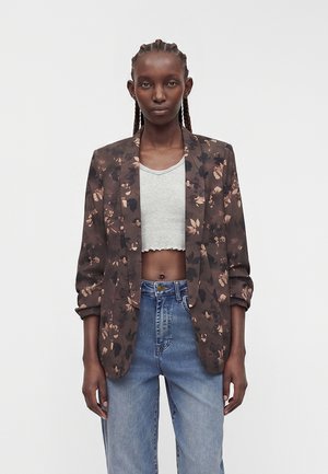 Femme aux cheveux tressés portant un blazer à fleurs sombre sur un top blanc court et un jean bleu taille haute, debout devant un fond uni.