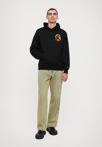 Sort hættejakke med en frontlomme, der har et orange astronautgrafik. Kombineret med lysegrønne stribede bukser og sorte sneakers.