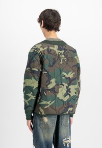 Persona con capelli corti castani che indossa una giacca trapuntata mimetica verde e marrone con la scritta "Alpha Industries" sulla schiena e jeans blu consumati.