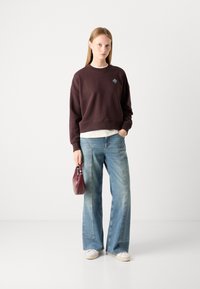 Sudadera marrón con un pequeño logo azul, combinada con vaqueros de pierna ancha de color azul claro, zapatillas blancas y un bolso burdeos.