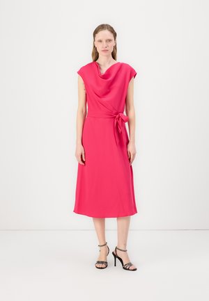 Robe rose, à mi-mollet, avec un col drapé, des manches courtes et une taille nouée. Tissu lisse avec une silhouette fluide. Talons fins noirs.