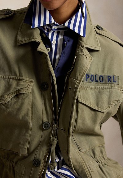 Közeli kép egy zöld munkakabátot viselő személyről, melyen a "POLO RL" feliratú címke látható, a csíkos kék-fehér, gombos ing fölött.