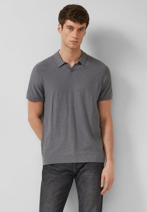 Junger Mann, der ein kurzärmeliges graues Strick-Poloshirt und dunkle Jeans trägt, steht vor einer schlichten weißen Wand.