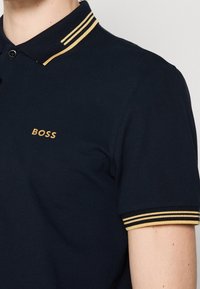 Polo bleu marine avec doubles rayures jaunes sur le col et les manches, et logo jaune "BOSS" sur la poitrine gauche.