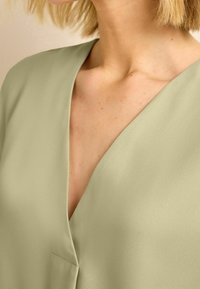 Sagegrüne Bluse mit V-Ausschnitt, aus leichtem, geschmeidigem Stoff und einem kleinen Fronttaschendetail.