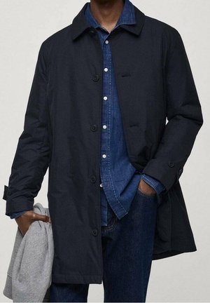 Homme portant un manteau noir boutonné par-dessus une chemise en denim bleu et un jean, tenant un pull gris plié dans une main.