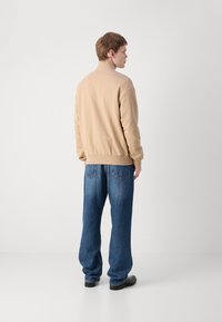 Felpa beige con colletto alto, vestibilità rilassata, polsini a coste e orlo dritto, abbinata a jeans in denim blu larghi con tasche posteriori.