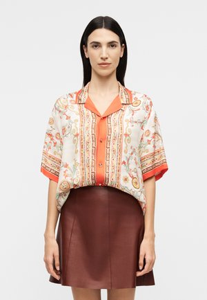 Γυναίκα με ίσια σκούρα μαλλιά, που φοράει πολύχρωμο πουκάμισο με κοντά μανίκια και καφέ δερμάτινη φούστα, στέκεται μπροστά σε ένα μονόχρωμο φόντο.