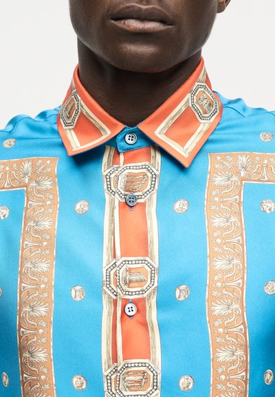 Detailný záber na osobu nosiacu modrú a oranžovú vzorovanú košeľu s gombíkmi, dekorovanú geometrickými a kvetinovými motívmi na golieri a prednej časti.