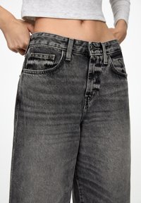 Jean gris taille haute avec une coupe décontractée, présentant un denim délavé, des poches avant et des accessoires métalliques à la taille et aux poches.