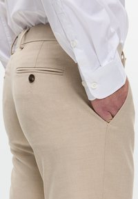 Pantaloni beige con una texture liscia, dotati di una tasca posteriore con bottone e di un cordino nascosto in vita, abbinati a una camicia bianca.