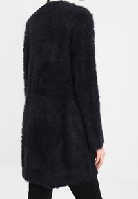 Lång, svart, fluffig cardigan bärs av en person, visad bakifrån, sträcker sig förbi höfterna med långa ärmar.