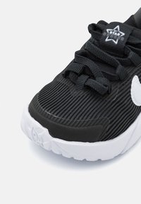 Chaussure de sport noire avec texture striée, accent de logo blanc, lacets plats et semelle extérieure blanche contrastante. Vue rapprochée de la zone des orteils.