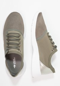 Chaussures de sport vert olive avec une tige en tissu texturé, des lacets plats, et une semelle en caoutchouc blanche ornée d'un accent argenté métallique au talon.