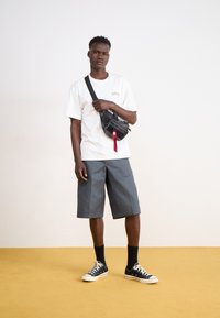 Hvit t-skjorte med kort erm og "Dickies"-merke, grå shorts til knærne, svart skulderveske, svarte sokker og svarte joggesko på gul gulv.