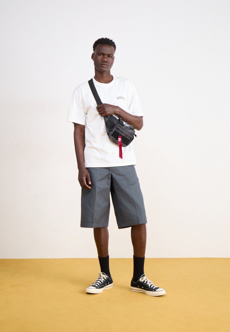 T-shirt bianca a maniche corte con logo "Dickies", pantaloni corti grigi fino al ginocchio, borsa crossbody nera, calzini neri e scarpe da ginnastica nere su un pavimento giallo.