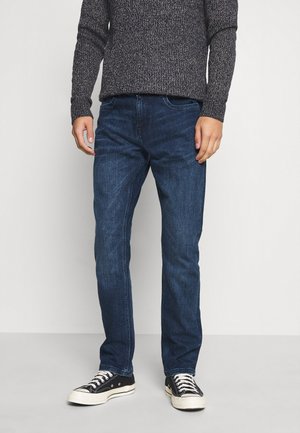 Dunkelblaue Jeans im geraden Schnitt, aus Denimstoff, kombiniert mit einem dunklen Pullover und schwarzen Low-Top-Sneakern mit weißen Schnürsenkeln.