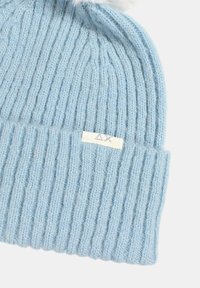 Beanie azzurro chiaro lavorato a maglia con una texture a coste, dotato di un'etichetta bianca con "ΔX" ricamata sull'orlo ripiegato e un fluffy pom-pom in cima.