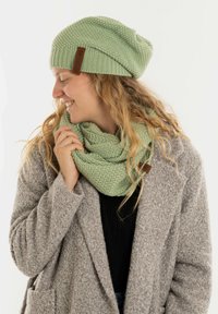 Groene gebreide beanie met een gestructureerd patroon en leren label, gecombineerd met een bijpassende groene gebreide sjaal, gedragen over een grijze jas.