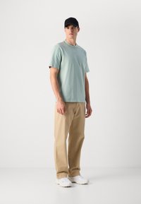 Jovem de pé, vestindo uma t-shirt verde clara, calças bege, sapatilhas brancas e um boné preto, contra um fundo branco liso.