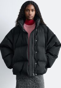Black puffer jacket with a high collar, silver zipper, and snap buttons; features quilted texture for insulation. Worn over a pink top.