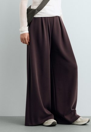 Personne portant un pantalon large marron foncé, une chemise blanche à manches longues, des baskets blanches et un sac noir en bandoulière, debout contre un mur gris.
