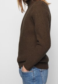 Brązowy sweter z golfem wykonany z dzianiny, o prążkowanej fakturze, dopasowanym kroju i długich rękawach, w połączeniu z jasnoniebieskimi dżinsami.