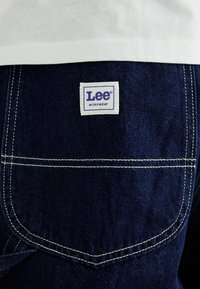 Tessuto denim blu scuro con un evidente motivo di cucitura bianco. Un'etichetta rettangolare presenta "Lee" in viola, sopra la parola "WORKWEAR".