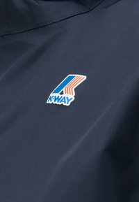 Ύφασμα σκούρου ναυτικού μπλε σακακιού με μπλε και πορτοκαλί μπαλώματα λογότυπου της εταιρείας K-Way στην περιοχή του στήθους.