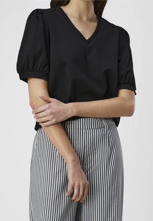 Mujer con blusa negra de manga abullonada con escote en V y pantalones de rayas verticales grises y blancos de talle alto, con los brazos cruzados al frente.