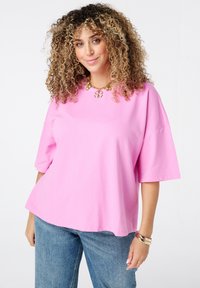 Vrouw met krullend haar, gekleed in een roze oversized shirt, blauwe jeans, gouden armband en ketting, glimlachend tegen een witte achtergrond.