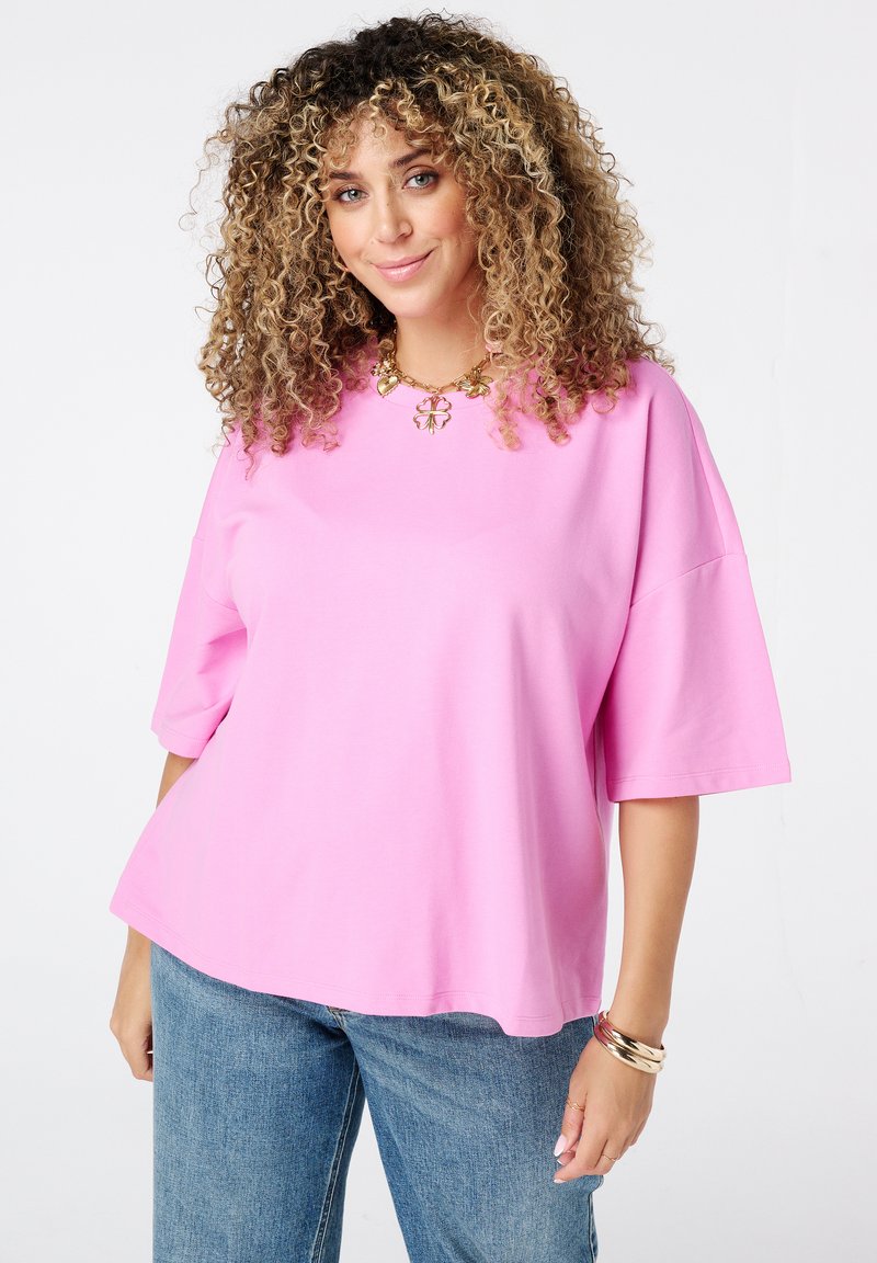 Vrouw met krullend haar, gekleed in een roze oversized shirt, blauwe jeans, gouden armband en ketting, glimlachend tegen een witte achtergrond.