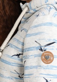 Sudadera gris claro con un patrón de rayas azules y gráficos de gaviotas negras. Incluye una placa de cuero marrón con detalles en relieve.